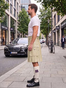 A man standing on a city sidewalk wearing a light beige kilt, white t-shirt, white knee-high socks, and black leather shoes, with a street scene and parked cars in the background.