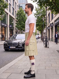 A man standing on a city sidewalk wearing a light beige kilt, white t-shirt, white knee-high socks, and black leather shoes, with a street scene and parked cars in the background.