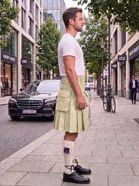 A man standing on a city sidewalk wearing a light olive green kilt, a white t-shirt, white socks, and black leather shoes, with a black Mercedes-Benz car and urban buildings in the background.
