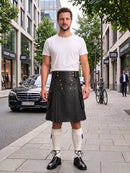 A man stands on a city sidewalk wearing a black leather kilt with gold buttons, a white t-shirt, white knee-high socks, and black leather shoes. The background shows a street with buildings, a parked black Mercedes, and a bicycle.