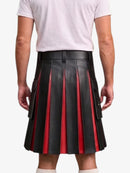 A man wearing a black pleated leather kilt with red vertical stripe accents, paired with a white t-shirt and white socks, shown from the back against a light gray background.