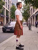 A man standing on a city sidewalk wearing a brown kilt with red pleats, a white t-shirt, white socks, and black leather shoes, with a street scene in the background.