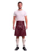 A man modeling a black faux-leather kilt with a matching black waist belt and pouch, paired with a white t-shirt, cream knee-high socks, and black lace-up shoes, against a plain white background.