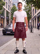 A man stands on a city sidewalk wearing a dark burgundy leather kilt with a silver buckle and strap, paired with a white t-shirt, white knee-high socks, and black leather shoes. A Mercedes-Benz car and city buildings are visible in the background.