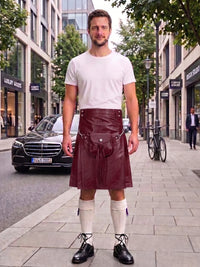 A man stands on a city sidewalk wearing a dark burgundy leather kilt with a silver buckle and strap, paired with a white t-shirt, white knee-high socks, and black leather shoes. A Mercedes-Benz car and city buildings are visible in the background.