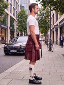 A man standing on a city sidewalk wearing a dark red leather kilt, a white t-shirt, white socks, and black leather shoes. A black Mercedes-Benz car is parked on the street behind him, with trees and storefronts in the background.