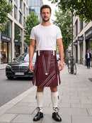 A man stands on a city sidewalk wearing a burgundy leather kilt with white contrast stitching, a white t-shirt, white knee-high socks, and black leather shoes. The background shows a blurred street scene with buildings, a parked black Mercedes, and a bicycle.