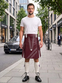 A man stands on a city sidewalk wearing a burgundy leather kilt with white contrast stitching, a white t-shirt, white knee-high socks, and black leather shoes. The background shows a blurred street scene with buildings, a parked black Mercedes, and a bicycle.
