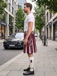 A man standing on a city sidewalk wearing a burgundy and white pleated kilt, white t-shirt, white knee-high socks, and black leather shoes, with a street scene and parked cars in the background.