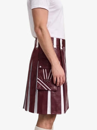 A side-profile view of a person wearing a pleated kilt in a deep burgundy faux leather with bold white vertical stripes and white drawstrings, paired with a white t-shirt against a plain white background.