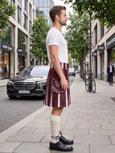 A man standing on a city sidewalk wearing a white t-shirt, a burgundy and white vertically striped kilt, cream knee-high socks, and black leather dress shoes. A black Mercedes-Benz car is parked on the street behind him.