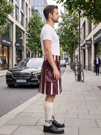 A man standing on a city sidewalk wearing a white t-shirt, a burgundy and white vertically striped kilt, cream knee-high socks, and black leather dress shoes. A black Mercedes-Benz car is parked on the street behind him.