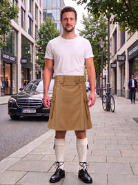 A man stands on a city sidewalk wearing a white crew-neck t-shirt and a tan pleated kilt with white knee-high socks and black leather shoes. The background shows a street scene with a Mercedes-Benz car, storefronts, and trees.