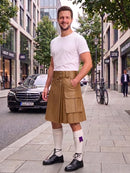 A man stands on a city sidewalk wearing a tan leather kilt with multiple flap pockets, a white t-shirt, white knee-high socks, and black leather shoes. A black Mercedes-Benz car is parked on the street behind him, with storefronts and trees in the background.