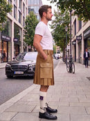 A man standing on a city sidewalk wearing a tan pleated kilt with a white t-shirt, white socks, and black leather shoes. The background shows a street with buildings, trees, and parked cars.