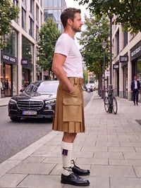 A man standing on a city sidewalk wearing a tan pleated kilt with a white t-shirt, white socks, and black leather shoes. The background shows a street with buildings, trees, and parked cars.