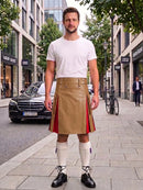 A man stands on a city sidewalk wearing a tan kilt with red side panels, a white t-shirt, white knee-high socks, and black shoes. Buildings, trees, and parked cars are visible in the background.