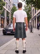 A man viewed from behind wearing a black leather kilt with lace-up back detailing, a white t-shirt, and white knee-high socks, standing on a city sidewalk.