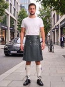 A man stands on a city sidewalk wearing a white crew-neck t-shirt and a dark gray pleated leather kilt, paired with white knee-high socks and black leather shoes. The background shows a street scene with a Mercedes-Benz car, storefronts, and trees.