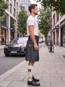 A man standing on a city sidewalk wearing a dark gray pleated kilt, a white t-shirt, white socks, and black leather shoes, with a street scene and parked cars in the background.