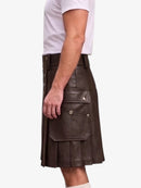 A man wearing a dark brown faux-leather kilt with pleated detailing and front cargo pockets, paired with a white t-shirt, shown from a side profile against a light gray background.