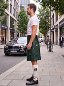 A man standing on a city sidewalk wearing a dark green leather kilt, white t-shirt, white knee-high socks, and black leather shoes. A black Mercedes-Benz car is parked on the street behind him, with trees and storefronts in the background.