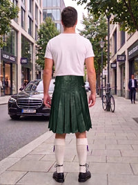 A man standing on a city sidewalk, viewed from behind, wearing a dark green pleated leather kilt, a white t-shirt, white knee-high socks, and black shoes. Buildings, trees, a parked black car, and a bicycle are visible in the background.