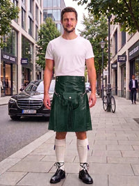 A man wearing a dark green leather kilt with a front pouch, a white t-shirt, white knee-high socks, and black leather shoes, standing on a city street sidewalk.