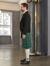 Man wearing a black jacket and green plaid kilt in an elegant interior setting.