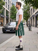 A man standing on a city sidewalk wearing a green and white striped kilt with a white V-neck t-shirt, white socks, and black leather shoes. A black Mercedes-Benz car and urban buildings are visible in the background.
