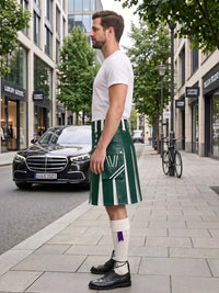 A man standing on a city sidewalk wearing a green and white striped kilt with a white V-neck t-shirt, white socks, and black leather shoes. A black Mercedes-Benz car and urban buildings are visible in the background.