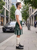 A man standing on a city sidewalk wearing a white short-sleeve t-shirt and a green kilt with white vertical stripes, paired with cream knee-high socks and black leather shoes. A black Mercedes-Benz car and a bicycle are visible in the background of the urban street scene.