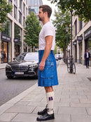 A man standing on a city sidewalk wearing a bright blue leather kilt, a white t-shirt, white socks, and black leather shoes. The background shows a street with a black Mercedes-Benz car, trees, and buildings.
