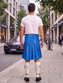 A man standing on a city sidewalk, viewed from behind, wearing a white t-shirt, a bright blue pleated faux-leather kilt, white knee-high socks, and black shoes. Buildings, trees, a parked black car, and a bicycle are visible in the background.