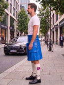 A man standing on a city sidewalk wearing a bright blue kilt with multiple flap pockets, a white t-shirt, white knee-high socks, and black shoes. A black Mercedes-Benz car is parked on the street behind him, with storefronts, trees, and a bicycle in the background.