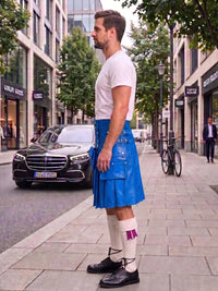 A man standing on a city sidewalk wearing a bright blue kilt with multiple flap pockets, a white t-shirt, white knee-high socks, and black shoes. A black Mercedes-Benz car is parked on the street behind him, with storefronts, trees, and a bicycle in the background.