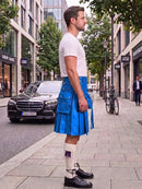 A man standing on a city sidewalk wearing a bright blue leather kilt, a white t-shirt, white socks, and black leather shoes. A black Mercedes-Benz car is parked on the street behind him, with buildings and trees in the background.