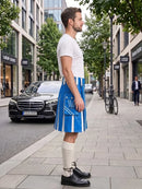 A man standing on a city sidewalk wearing a white short-sleeve T-shirt, a bright blue and white vertically striped kilt with front pockets, cream knee-high socks, and black leather shoes. A black Mercedes-Benz car and urban buildings are visible in the background.