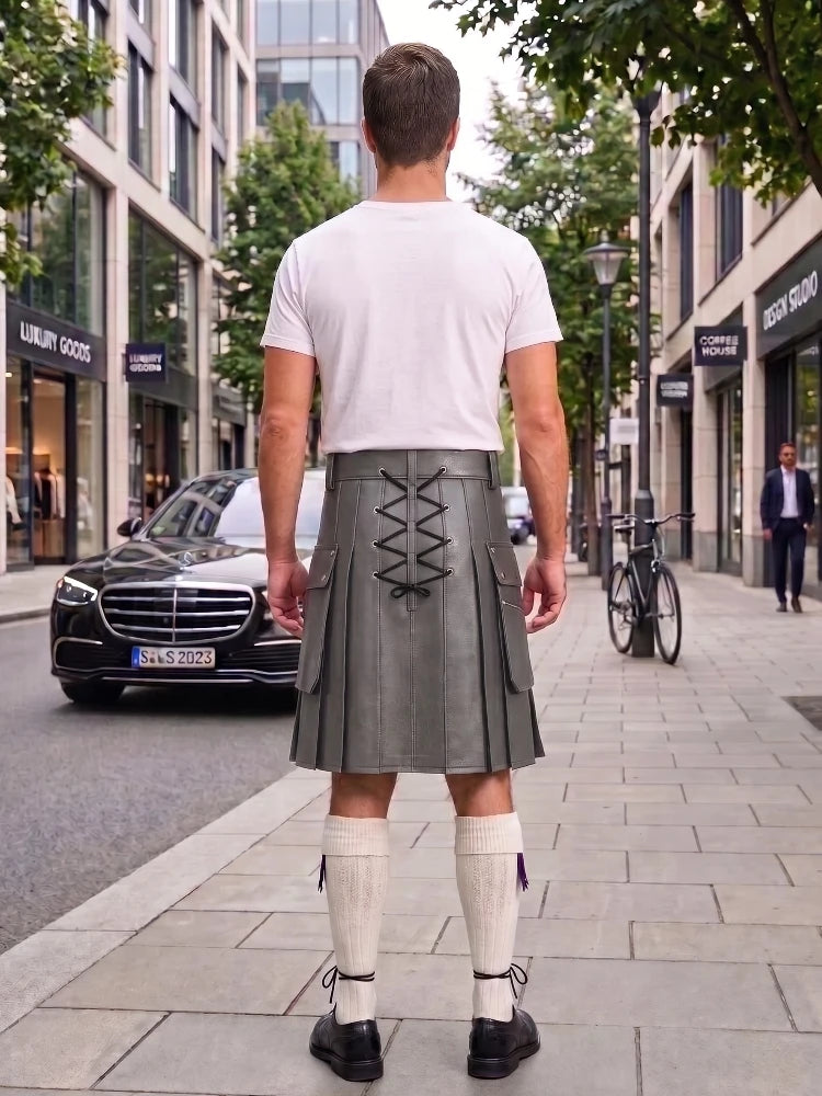 A man standing on a city sidewalk, viewed from behind, wearing a white t-shirt, a gray leather kilt with lace-up back detailing, white knee-high socks, and black shoes.