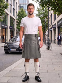 A man stands on a city sidewalk wearing a white short-sleeve t-shirt and a gray pleated leather kilt with silver hardware, paired with white knee-high socks and black leather shoes. Buildings, trees, and a parked black car are visible in the background.