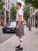 A man standing on a city sidewalk wearing a gray kilt, white t-shirt, white socks with purple accents, and black shoes. A black Mercedes-Benz car is parked behind him on the street, with trees and storefronts in the background.