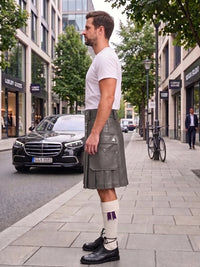 A man standing on a city sidewalk wearing a gray kilt, white t-shirt, white socks with purple accents, and black shoes. A black Mercedes-Benz car is parked behind him on the street, with trees and storefronts in the background.