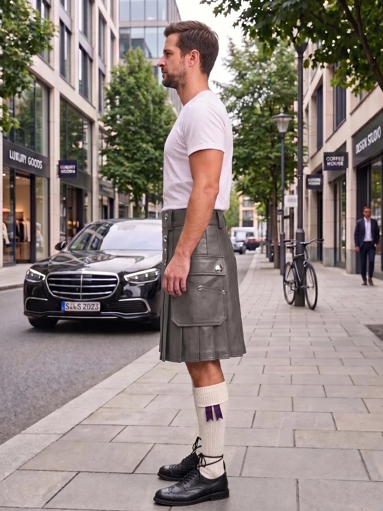 A man standing on a city sidewalk wearing a gray kilt, white t-shirt, white socks with purple accents, and black shoes. A black Mercedes-Benz car is parked behind him on the street, with trees and storefronts in the background.