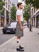 A man standing on a city sidewalk wearing a white t-shirt, a gray pleated kilt with front pockets, white socks with purple stripes, and black leather oxford shoes. A black Mercedes-Benz car and a bicycle are parked on the street behind him.