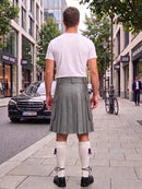 A man standing on a city sidewalk, viewed from behind, wearing a white t-shirt, a pleated gray kilt, white knee-high socks, and black shoes. Storefronts, trees, and a parked black car are visible in the background.