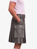 A person wearing a dark gray faux-leather pleated kilt with a matching belt and silver buckle, paired with a light pink t-shirt, shown from the side against a plain white background.