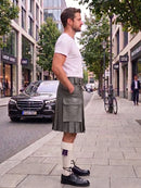 A man wearing a gray kilt, white t-shirt, white socks with a purple ribbon, and black leather shoes stands on a city sidewalk next to a street with parked cars and trees.