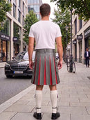 A man viewed from behind wearing a gray pleated kilt with red center panels, a white t-shirt, white knee-high socks, and black shoes, standing on a city sidewalk.