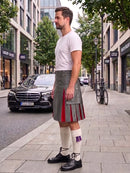 A man standing on a city sidewalk wearing a gray kilt with red pleated accents, a white t-shirt, white knee-high socks, and black shoes. A street, parked cars, and buildings are visible in the background.
