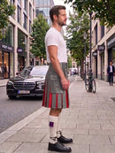 A man standing on a city sidewalk wearing a gray kilt with red pleated detailing, a white t-shirt, white socks, and black leather shoes, with a street scene in the background.